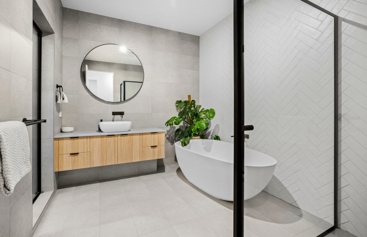 Modern bathroom with gray walls, wood vanity, round mirror, white tub, and glass shower. — Kolora Projects in Robina, QLD