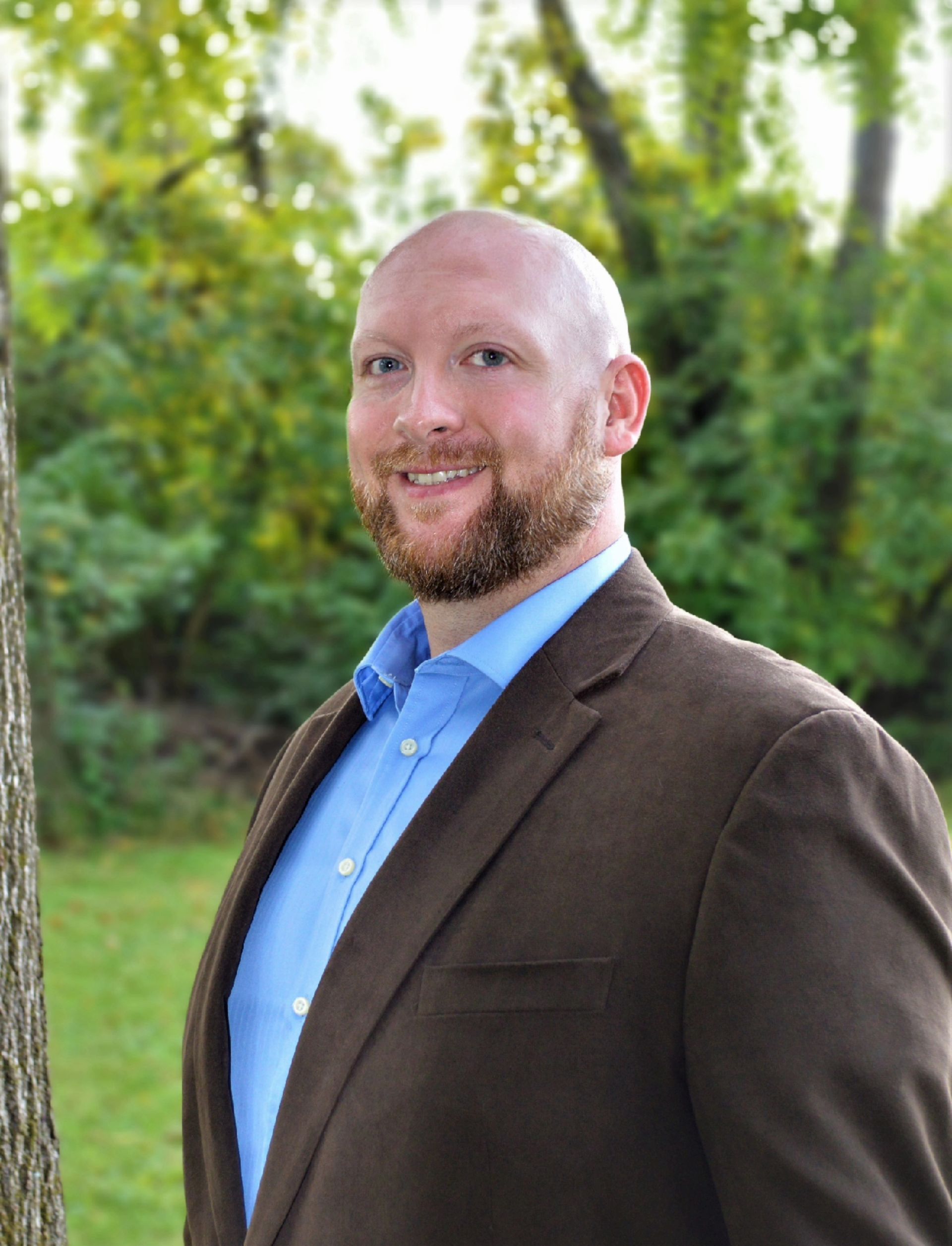 A man in a suit and blue shirt is standing in front of a tree.