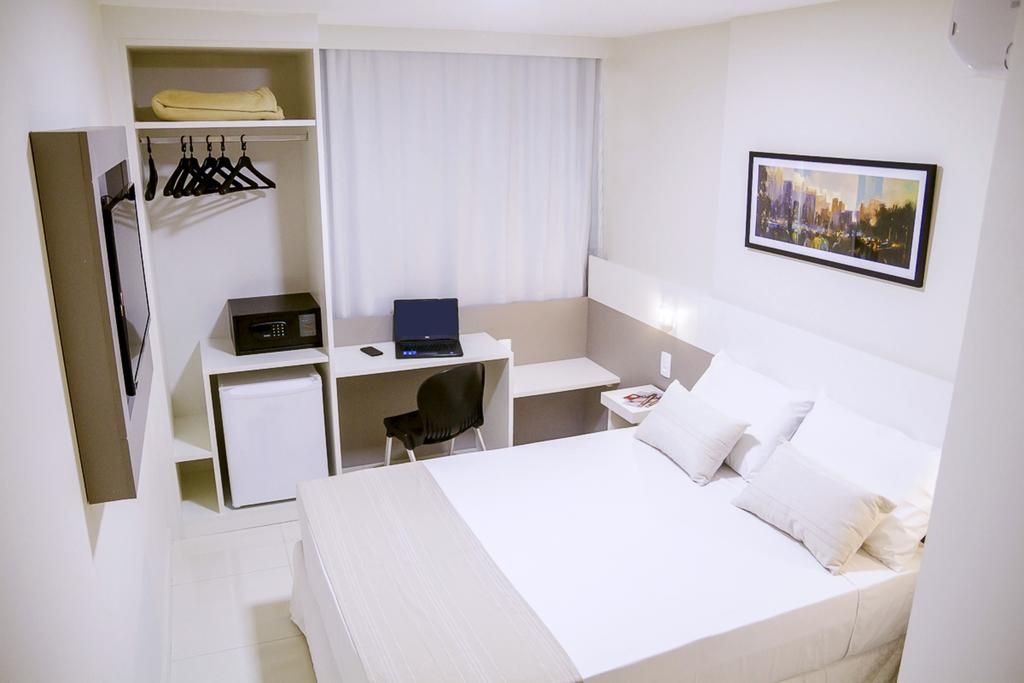 Quarto de hotel com cama, escrivaninha, armário e obras de arte. Esquema de cores branco e bege.