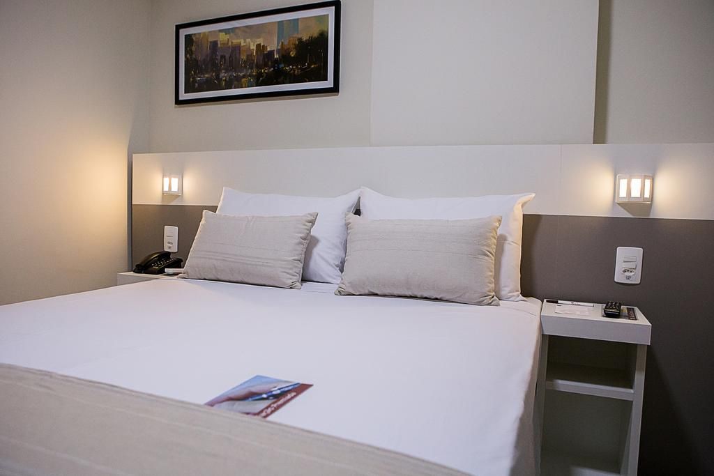 Quarto de hotel com cama arrumada, travesseiros, luminárias, mesa de cabeceira e quadros.