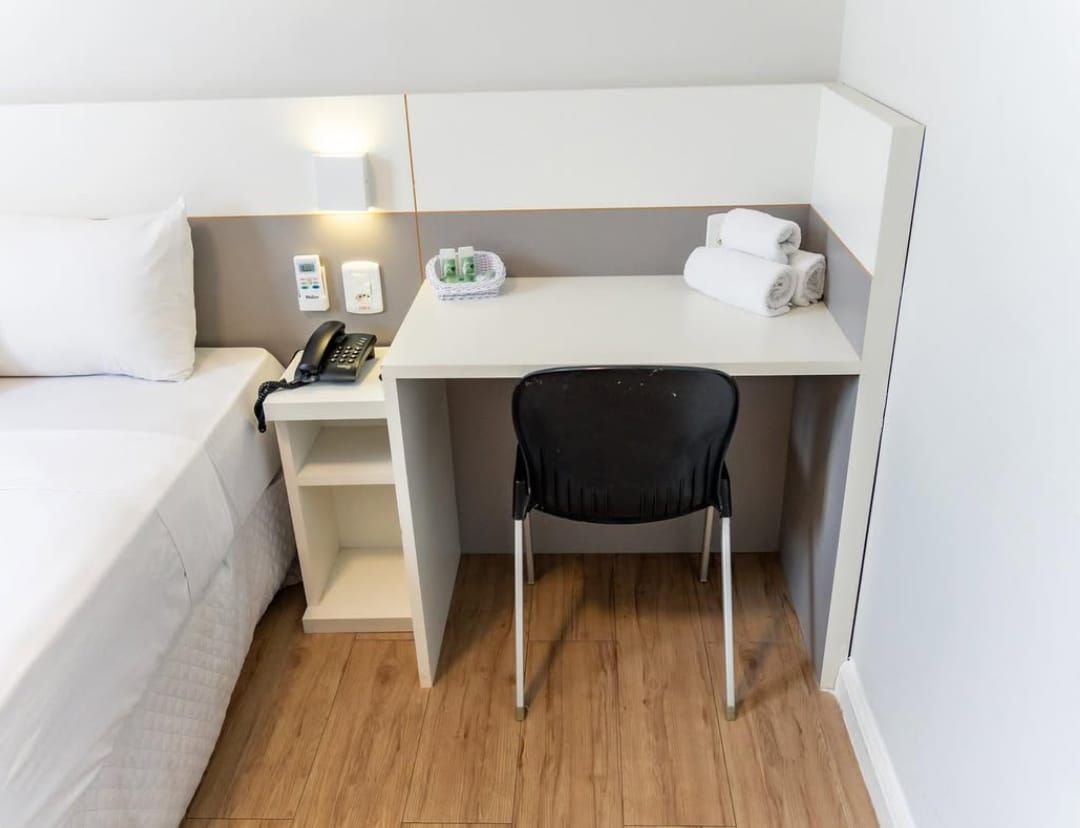 Recepção do hotel: mesa branca, cadeira preta, telefone, toalhas e uma cama com travesseiro branco.