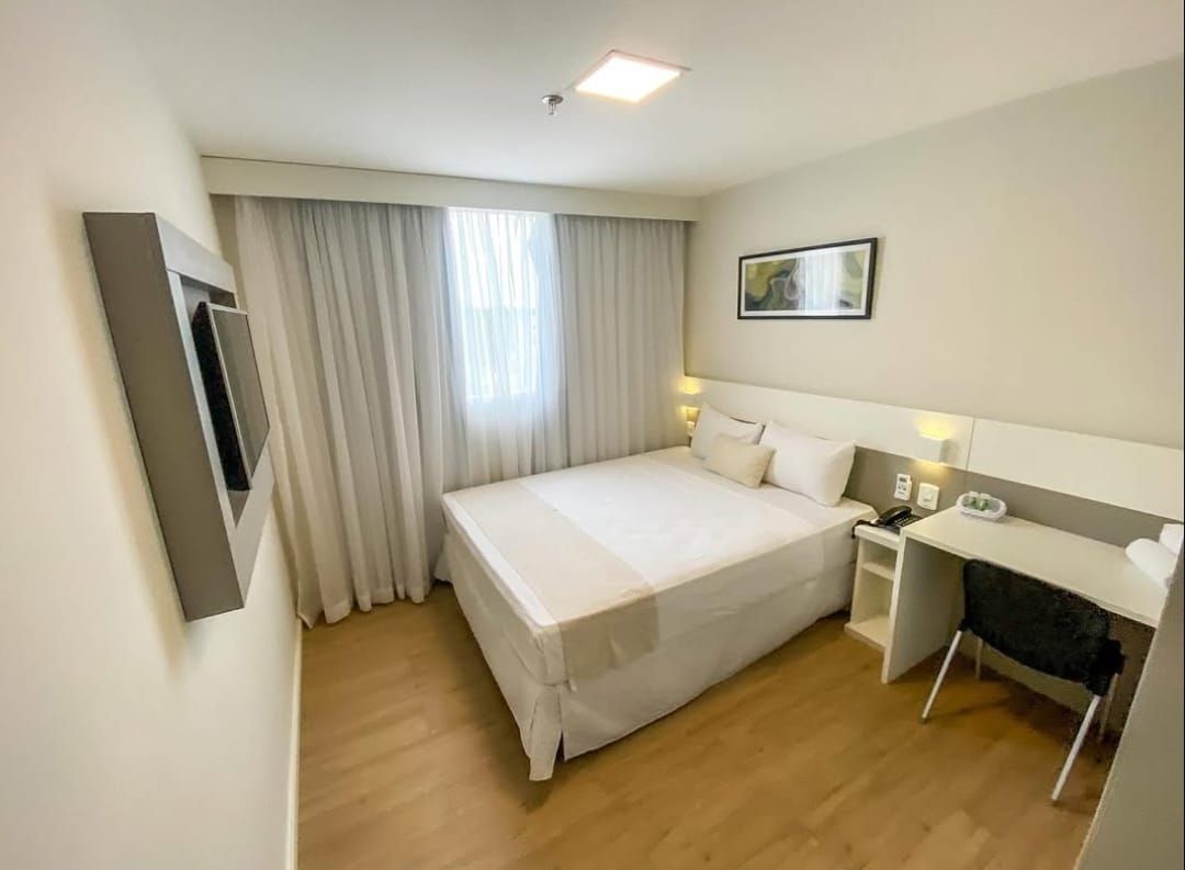 Quarto de hotel com cama, escrivaninha, TV e janela com cortinas claras.