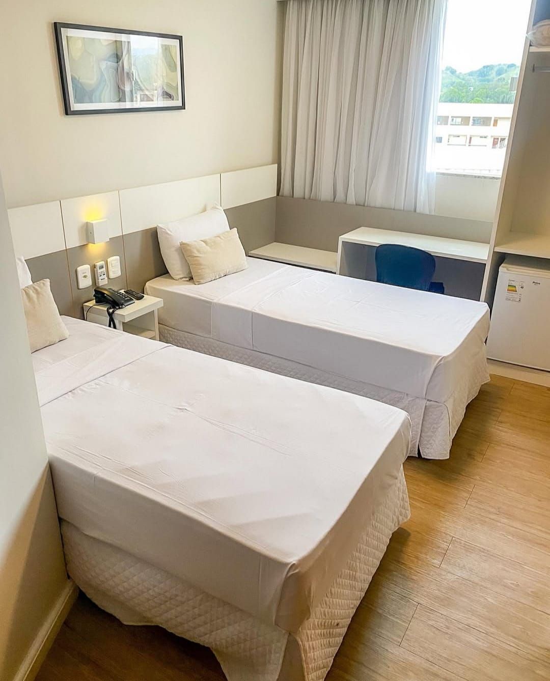 Quarto de hotel com duas camas de solteiro, camas brancas, escrivaninha e geladeira pequena.