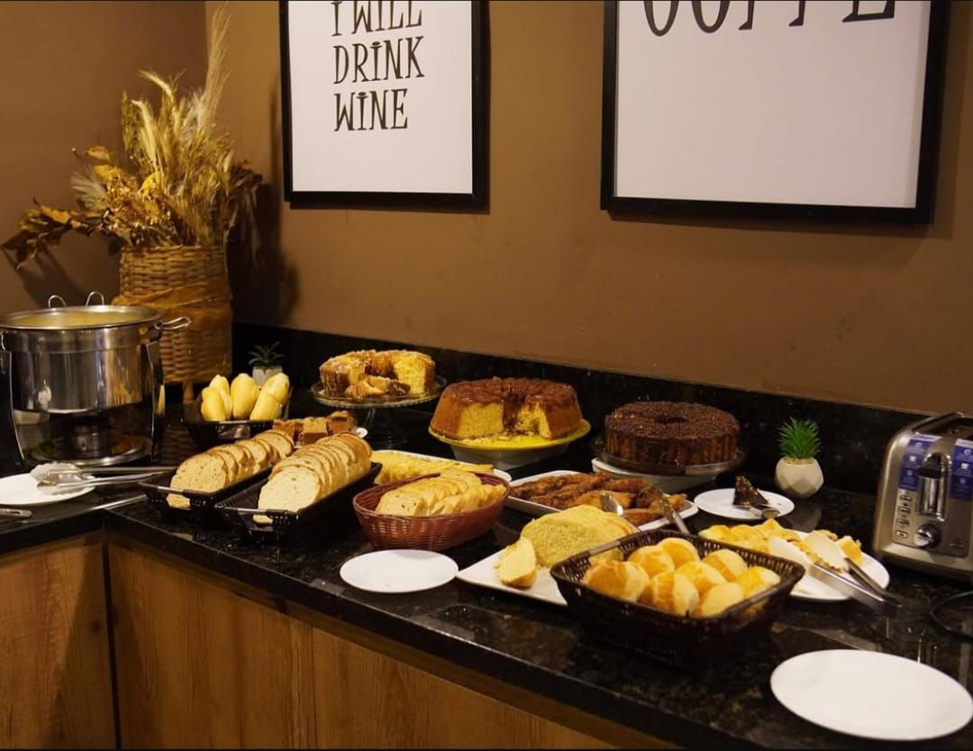 Buffet de café da manhã com diversos bolos, pães e doces em um balcão sob placas emolduradas.
