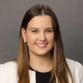 Headshot of a smiling person with long brown hair, wearing a white blazer and pearl necklace on a gray background