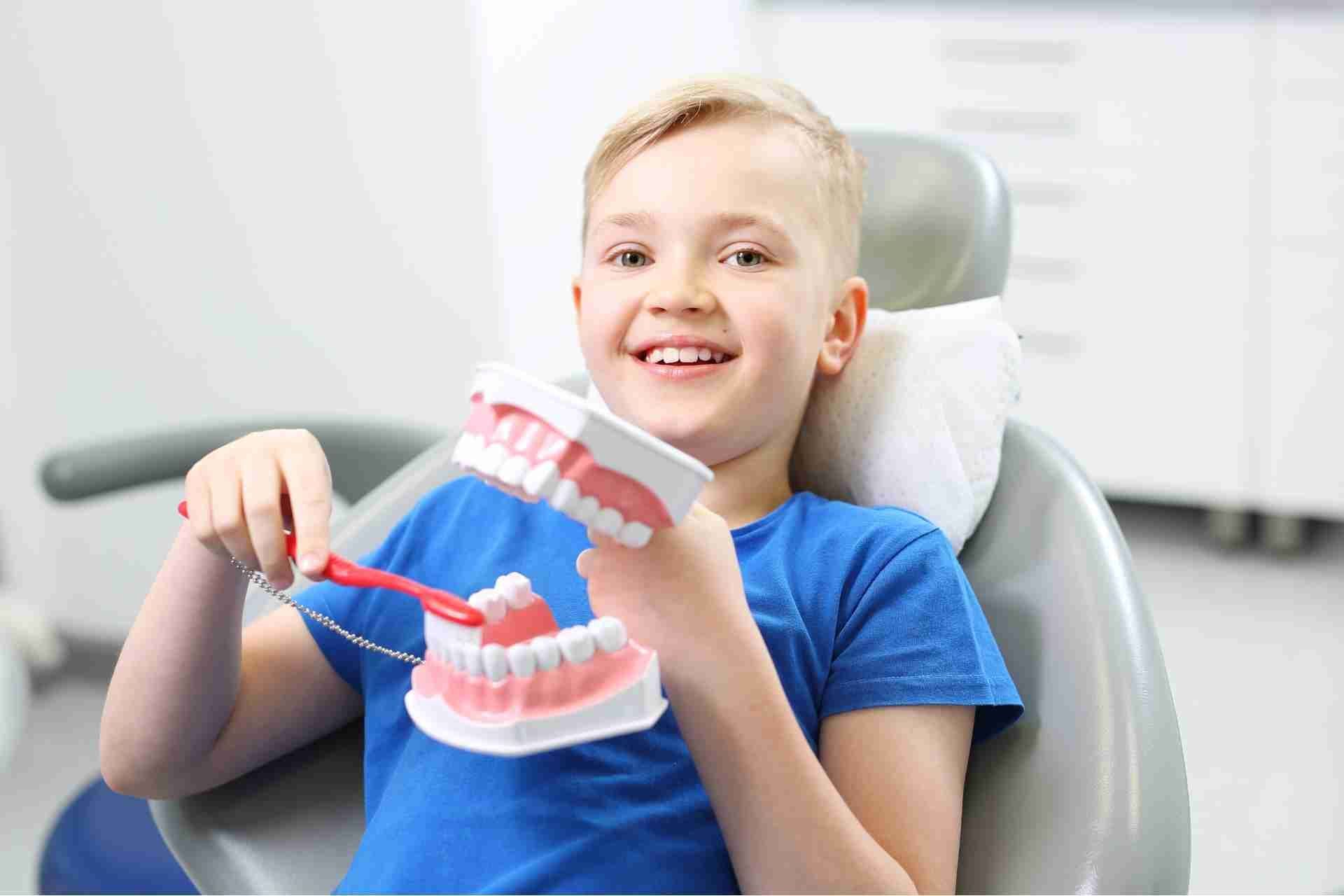 Child smiling in a dental chair, holding a model of teeth with a toothbrush