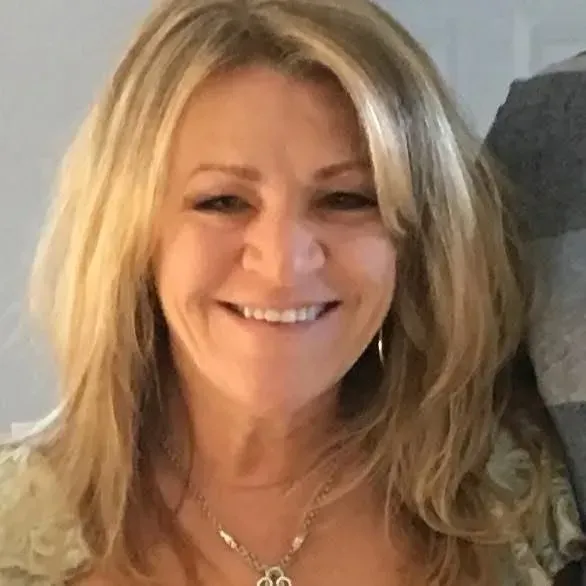 Smiling blonde woman indoors, wearing a necklace and light-colored top.