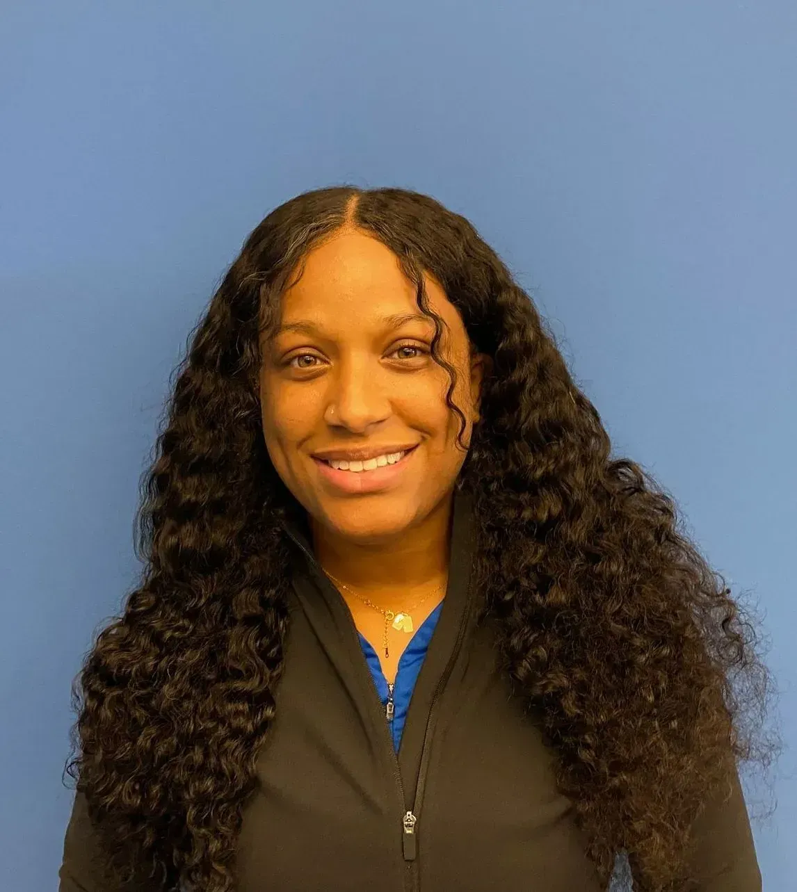 Smiling person with long curly hair in a black jacket against a blue background
