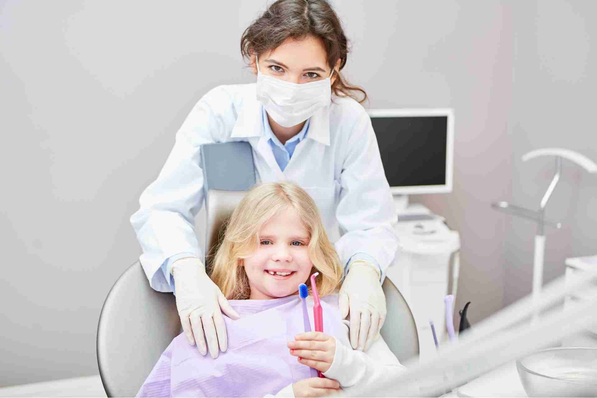 Masked dentist with a young patient in a dental office, smiling with dental tools visible.