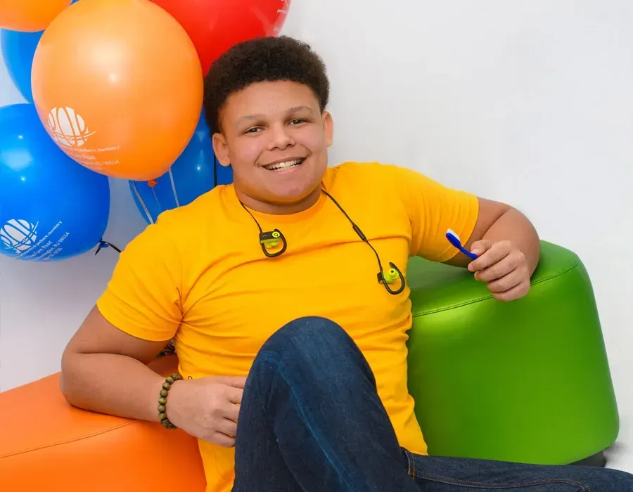 Smiling person in a yellow shirt sitting on colorful beanbag chairs with orange and blue balloons nearby