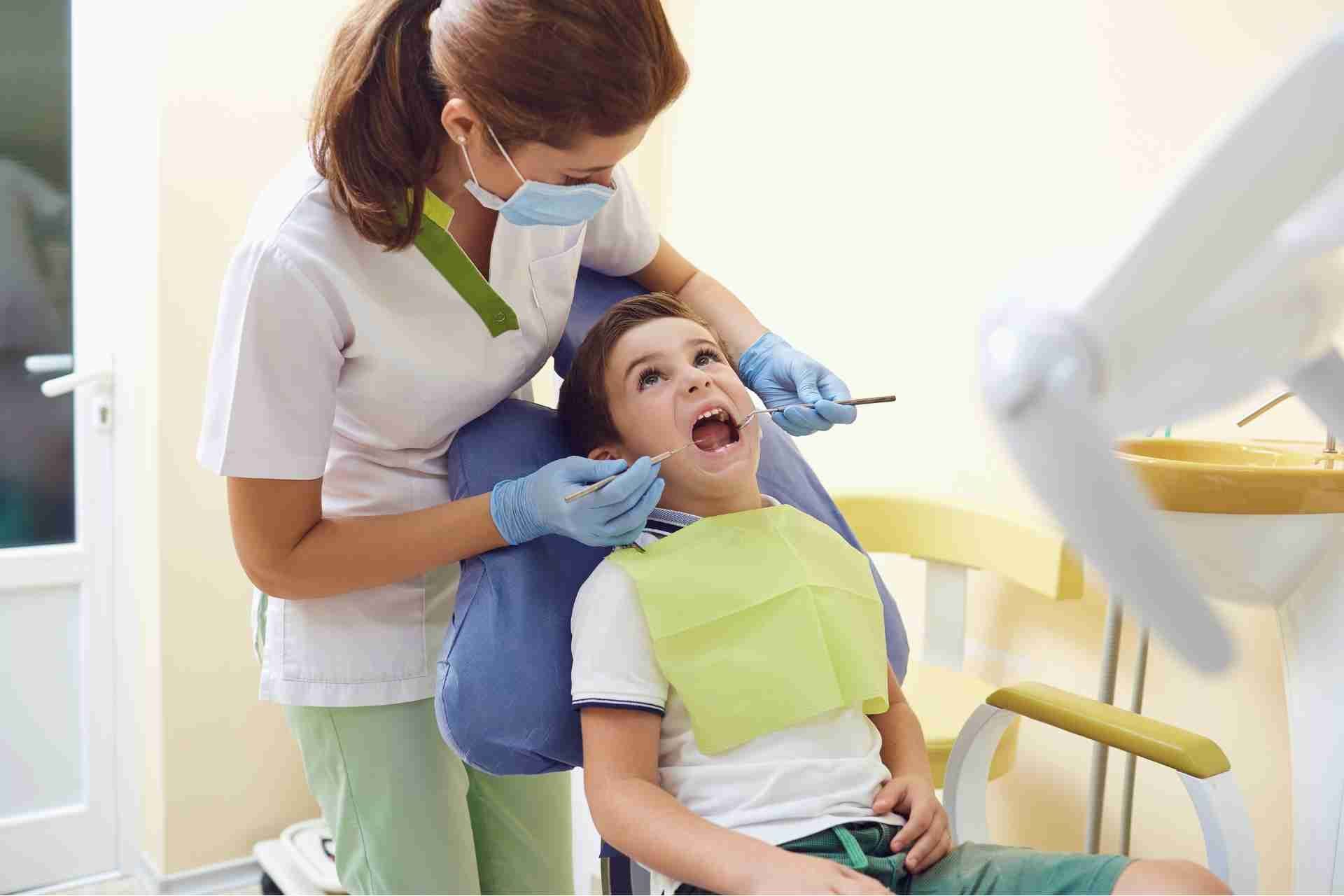 Dentist examining a young patient in a dental chair, with a blue bib and gloves in a bright clinic.