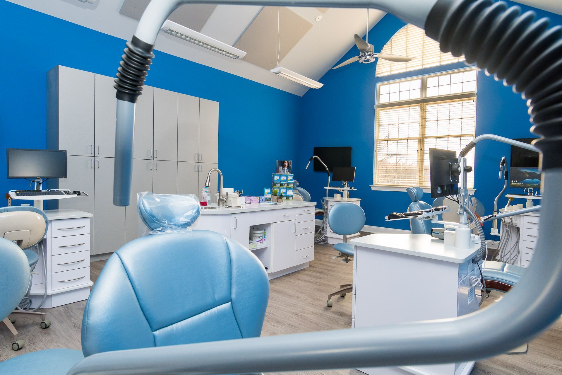 Bright blue dental clinic with chairs, desks, monitors, and large overhead ventilation hoses