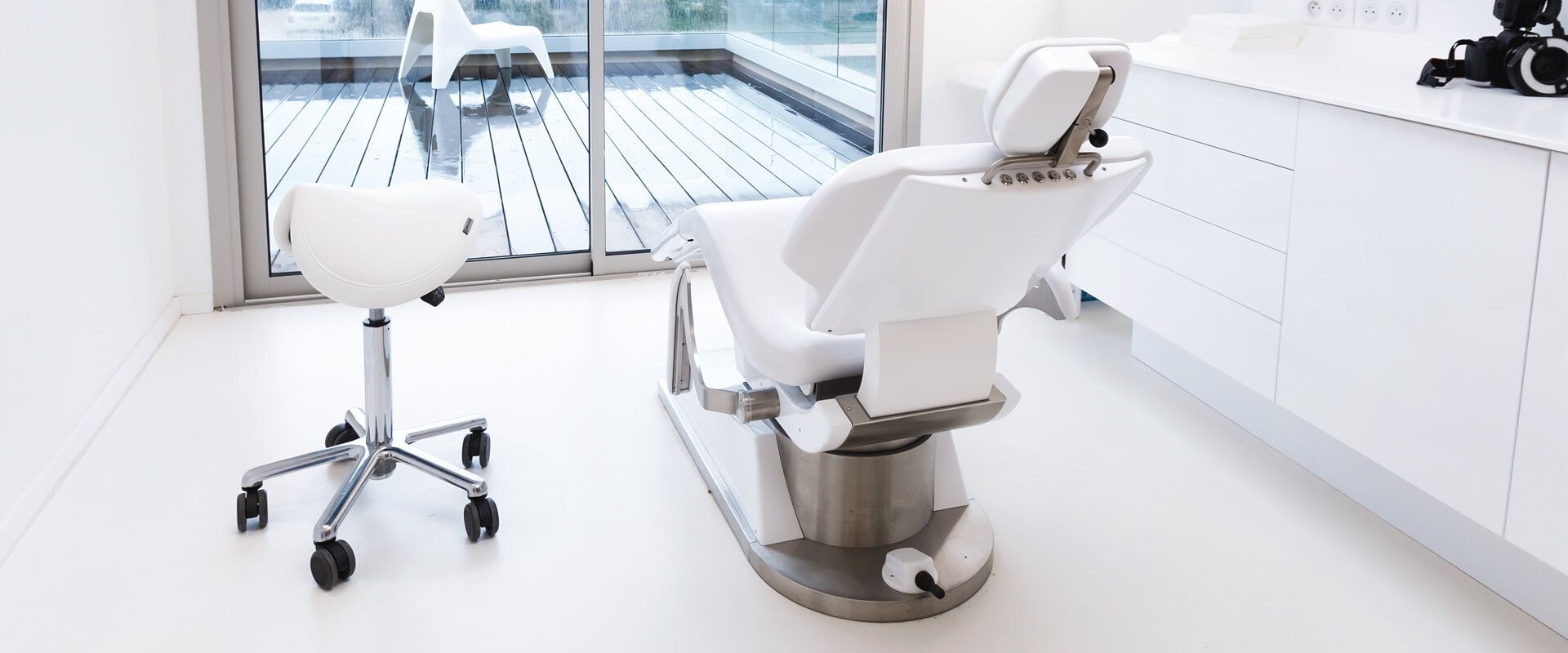 Bright dental exam room with white chair, stool, and cabinets by a window.