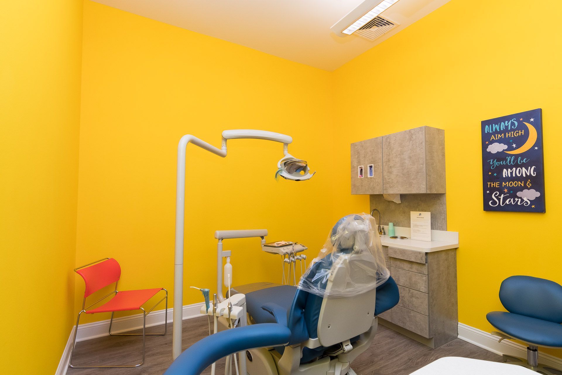 Bright yellow dental exam room with chair, overhead light, sink, and colorful waiting chairs