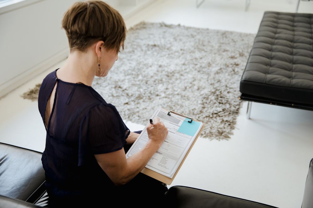 Person writing on a clipboard while seated on a black sofa in a bright living room.