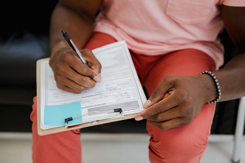 Person writing on a clipboard while seated, wearing coral pants and a pink shirt.