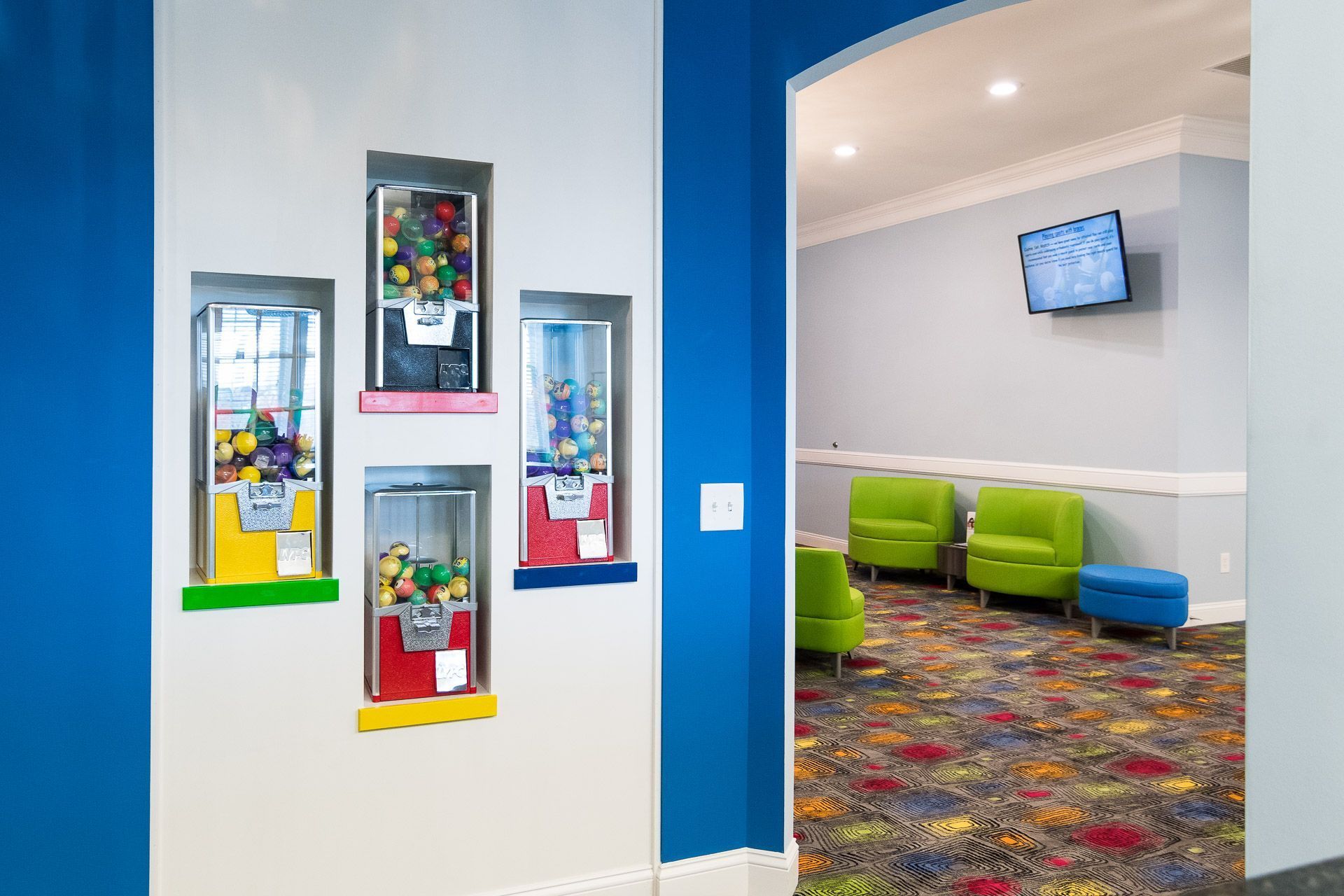 Colorful hallway with framed wall art, opening into a lounge with green chairs and a wall-mounted TV