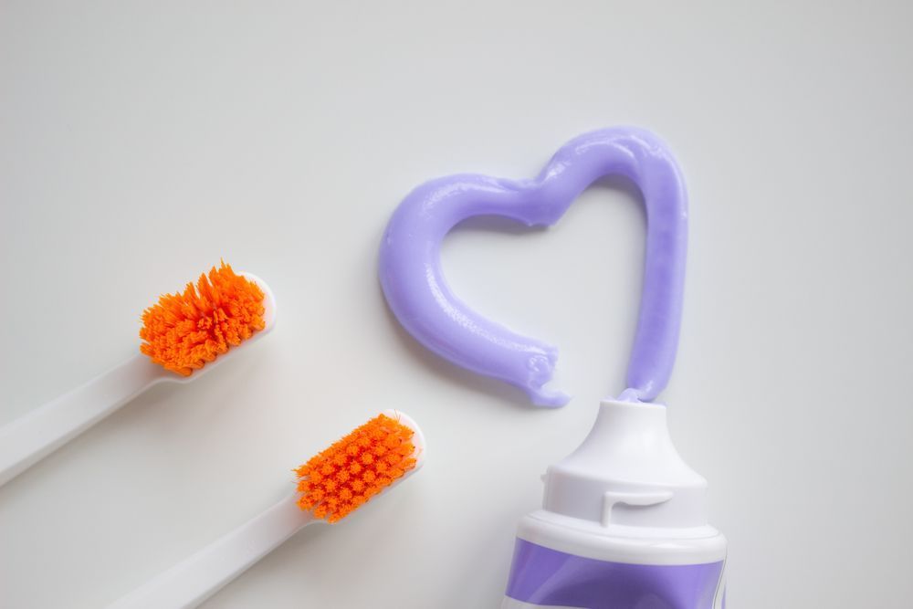 Purple toothpaste forming a heart above orange toothbrush bristles on a white background