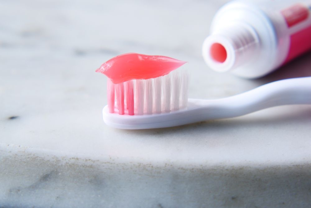 Pink toothpaste on a white toothbrush beside an open tube on a marble countertop