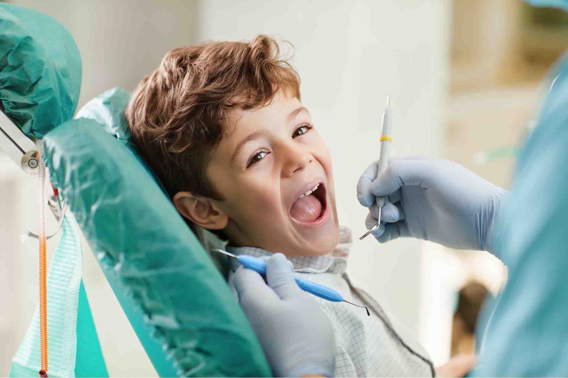 Child at dentist, smiling with mouth open during dental exam, gloved hand holding a tool nearby