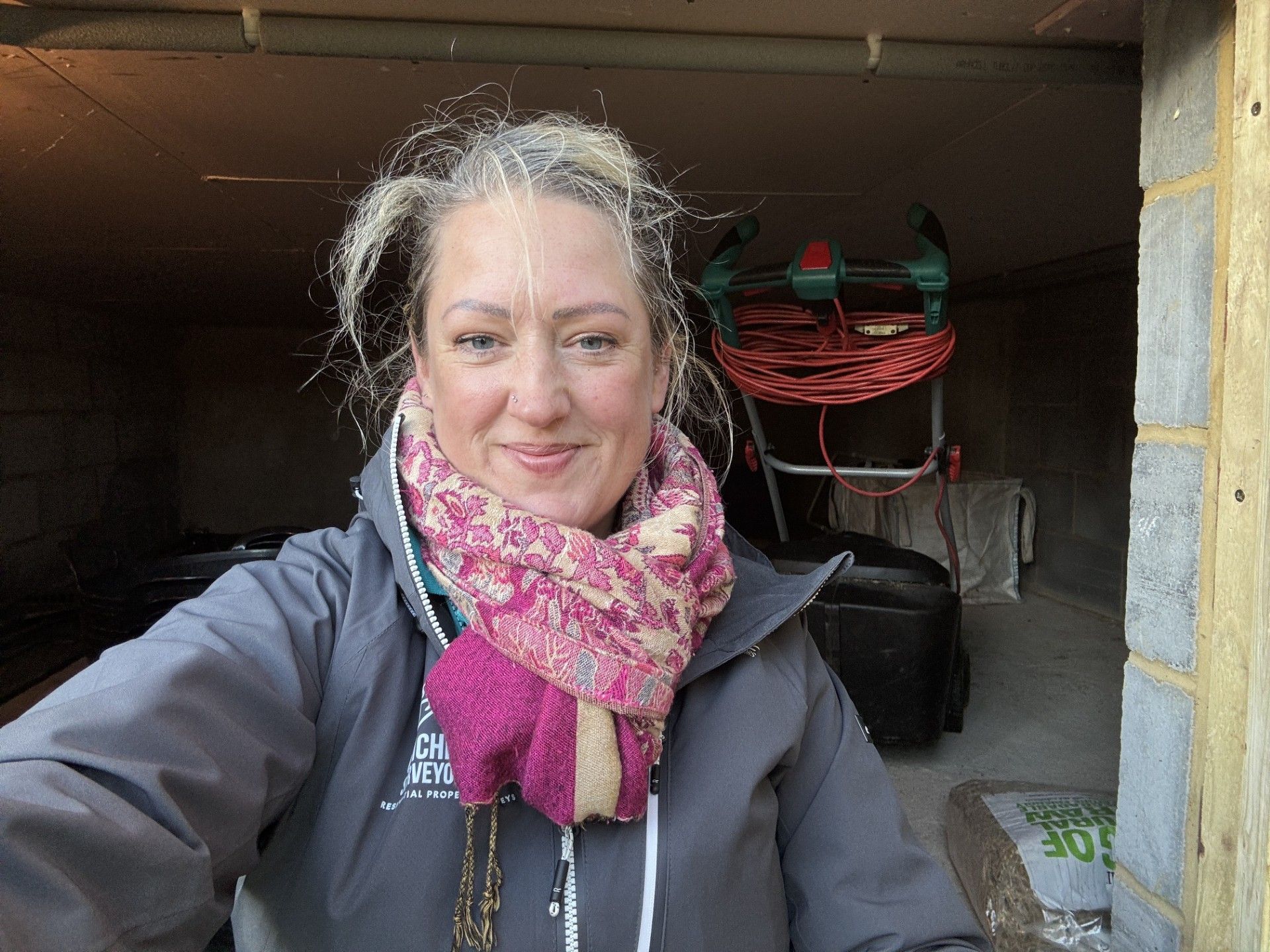 Photograph of Rebecca Jenkins wearing a pink scarf and grey jacket smiles, standing in a storage area.