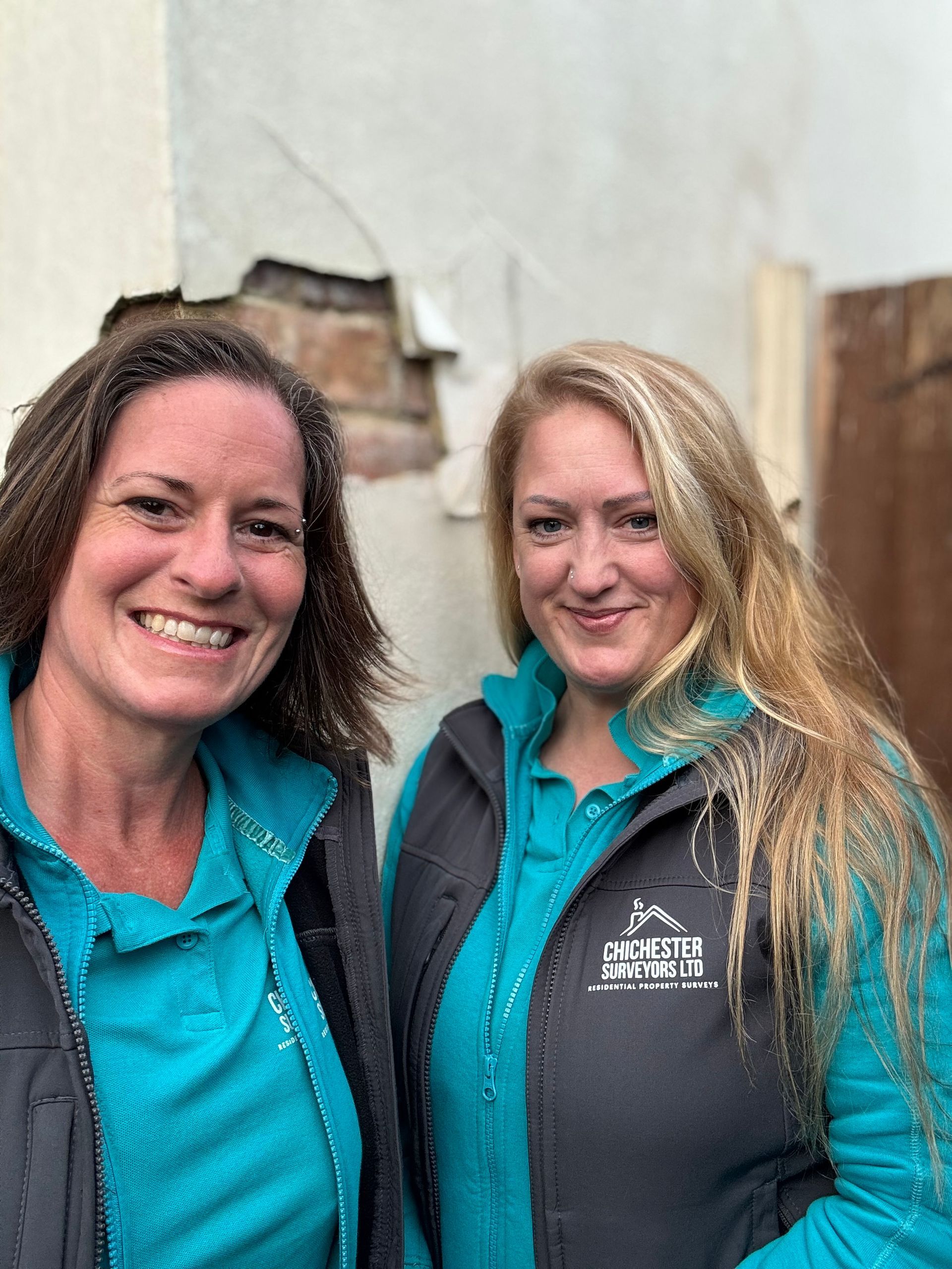 Two smiling people in teal jackets standing indoors against a damaged rendered wall.