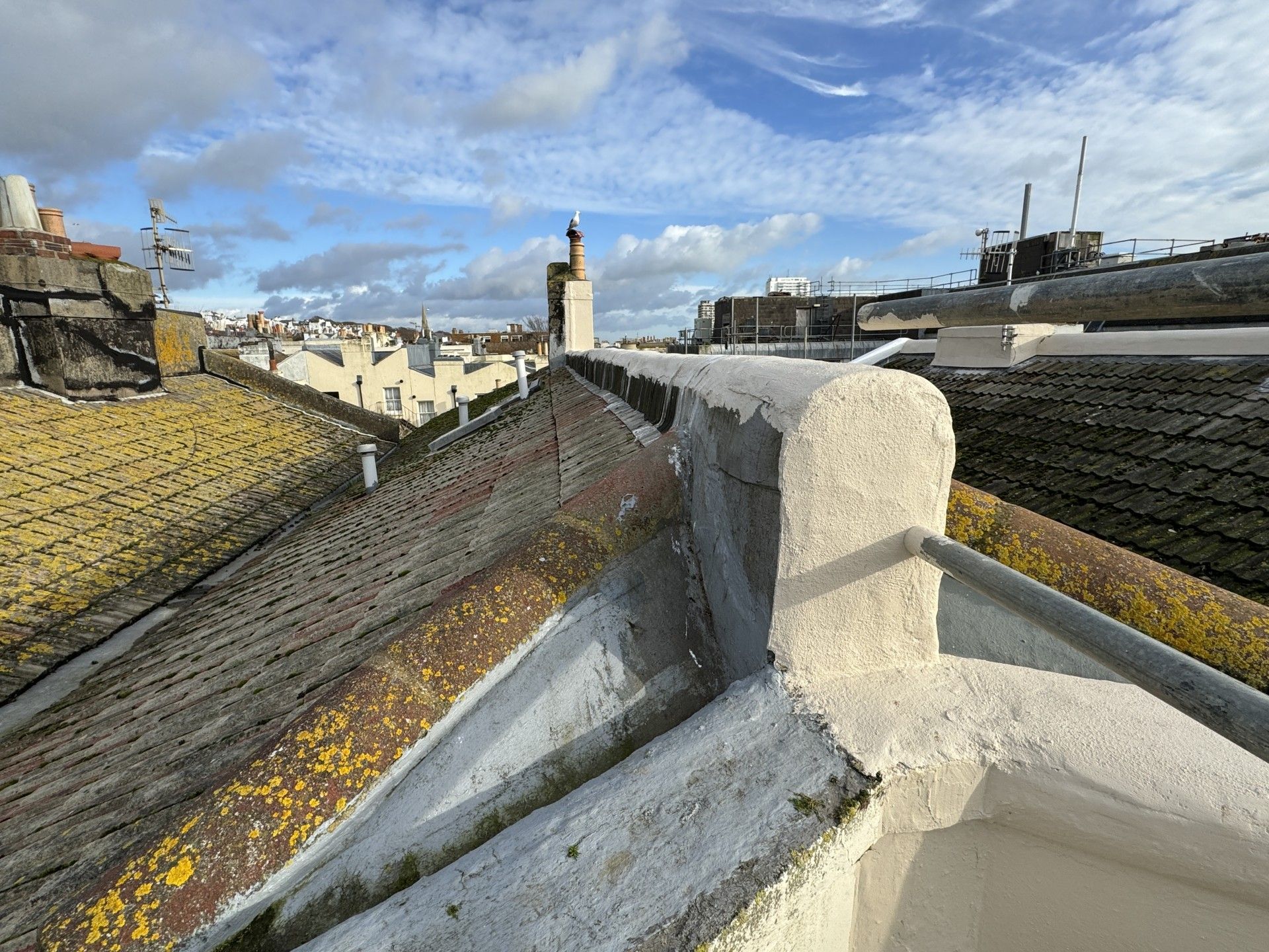 Rooftop view over a town with chimneys, tiled roofs, and a stone parapet under a cloudy sky
