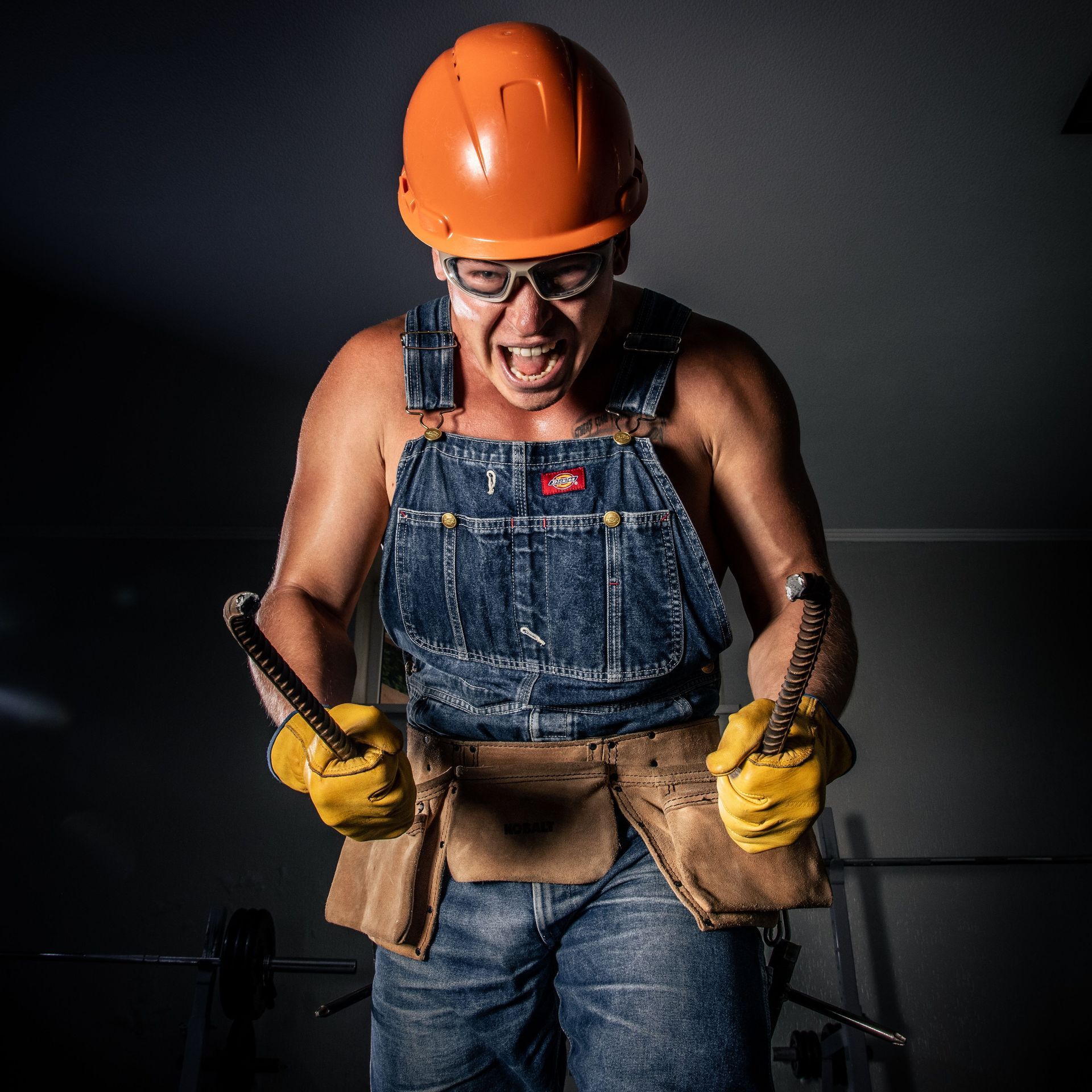 Worker in orange hard hat and denim overalls shouting while gripping tools against a dark background