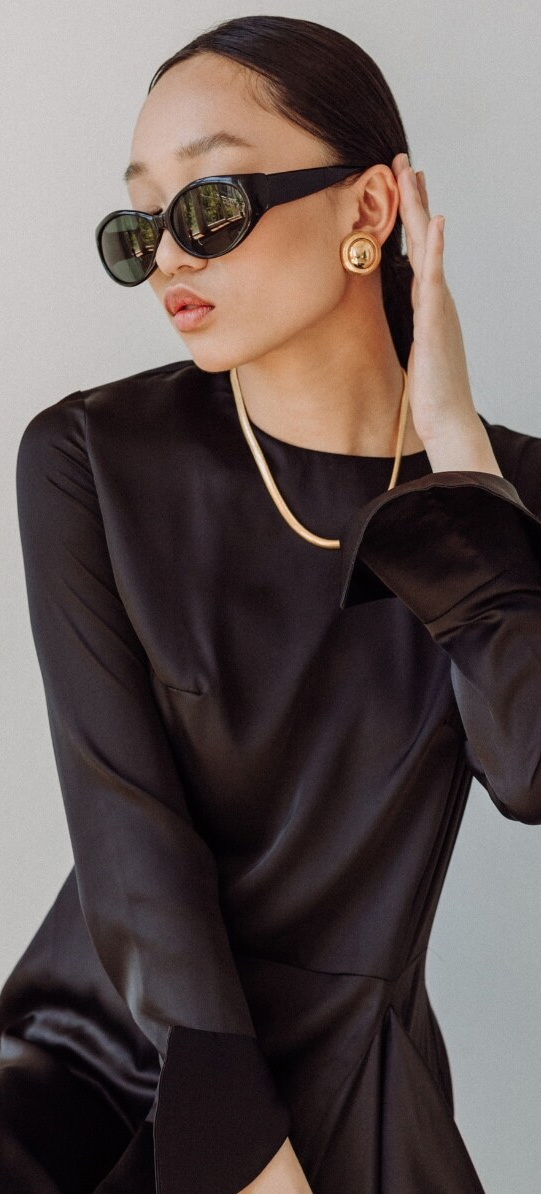 Persona vestita di nero, con occhiali da sole e gioielli d'oro, in posa su uno sfondo chiaro.