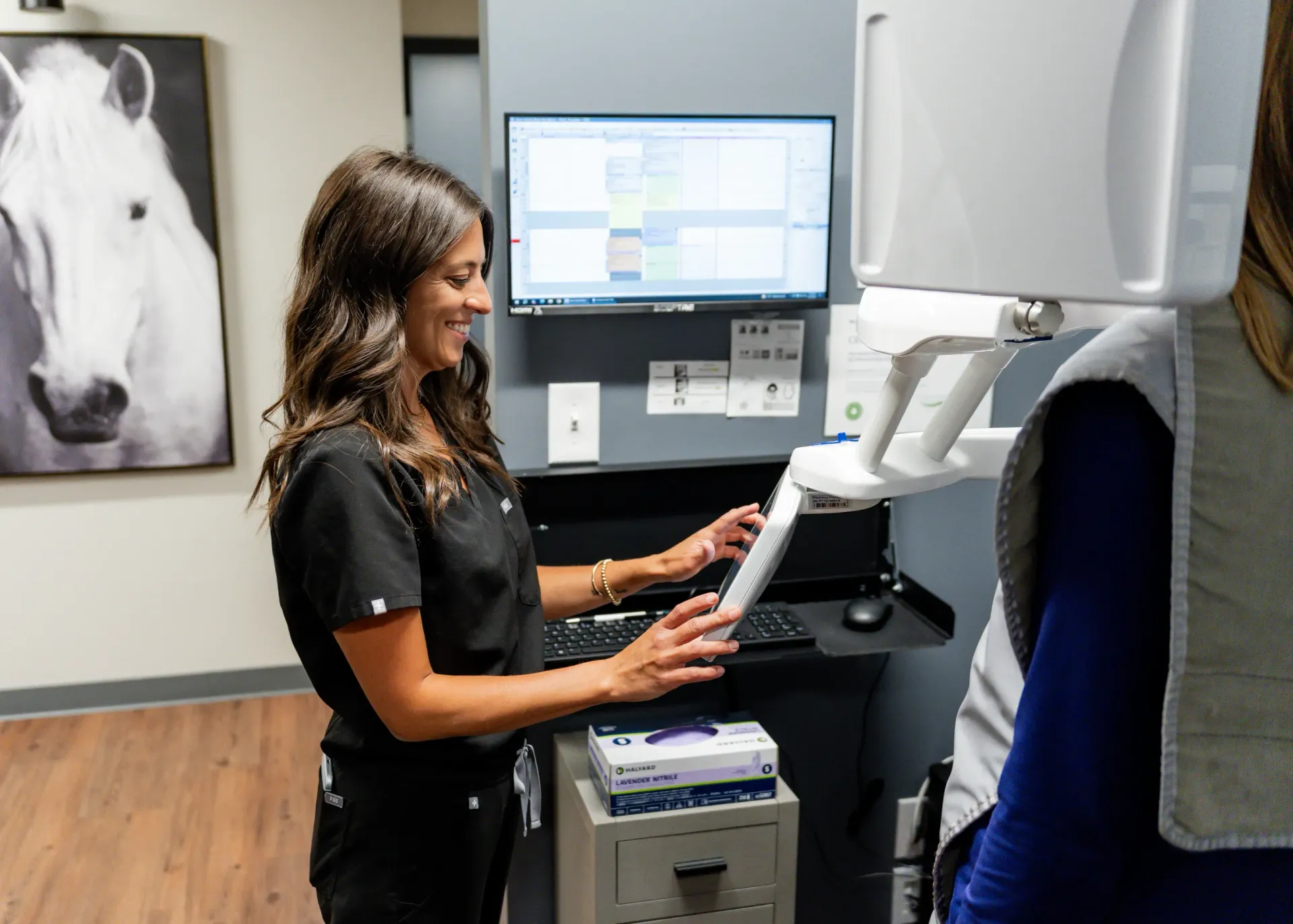 Dental assistant using a tablet near a patient, looking at a computer screen in a clinic setting.