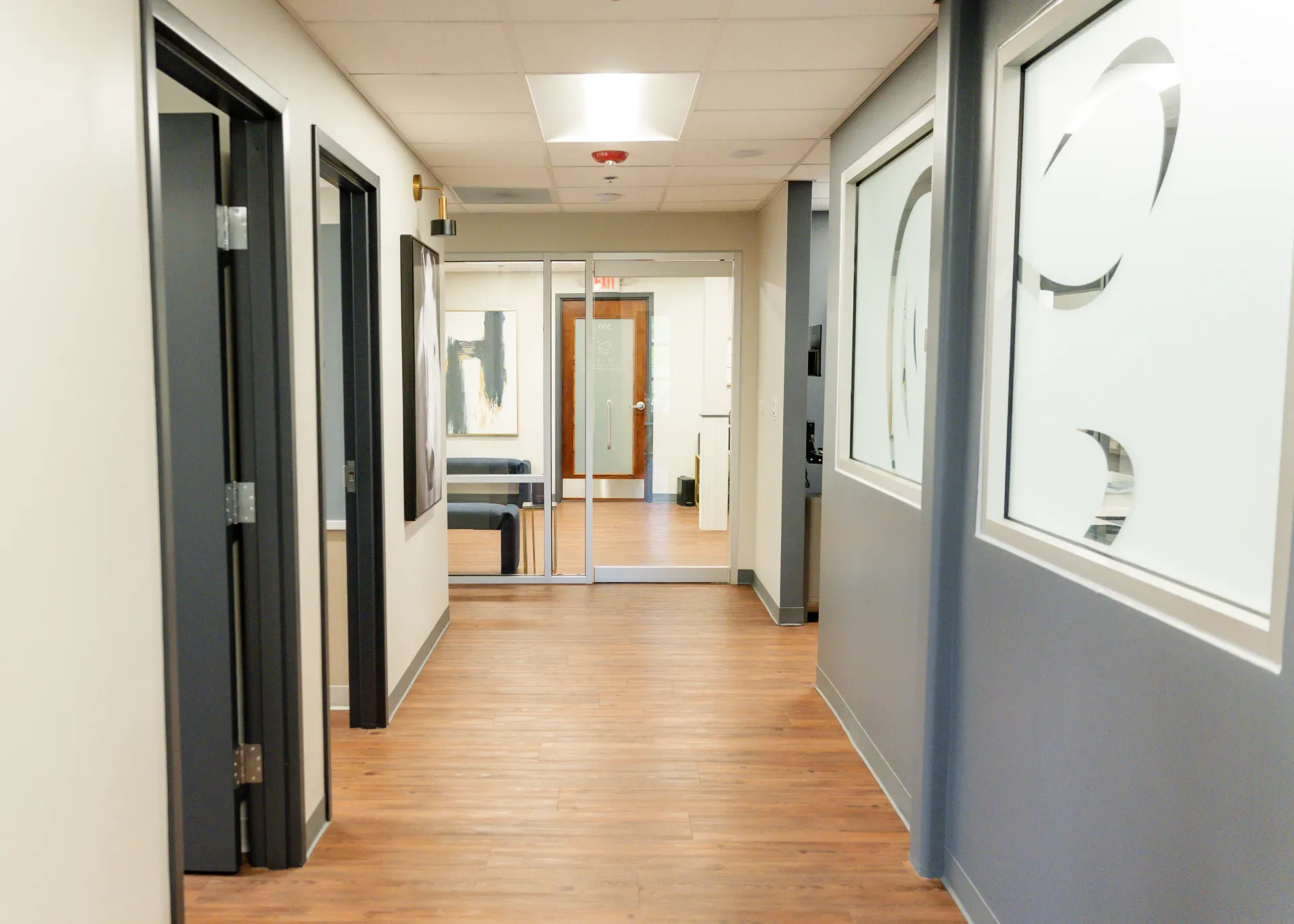 Hallway in a medical office with doors on the left, windows on the right, and a door at the end.