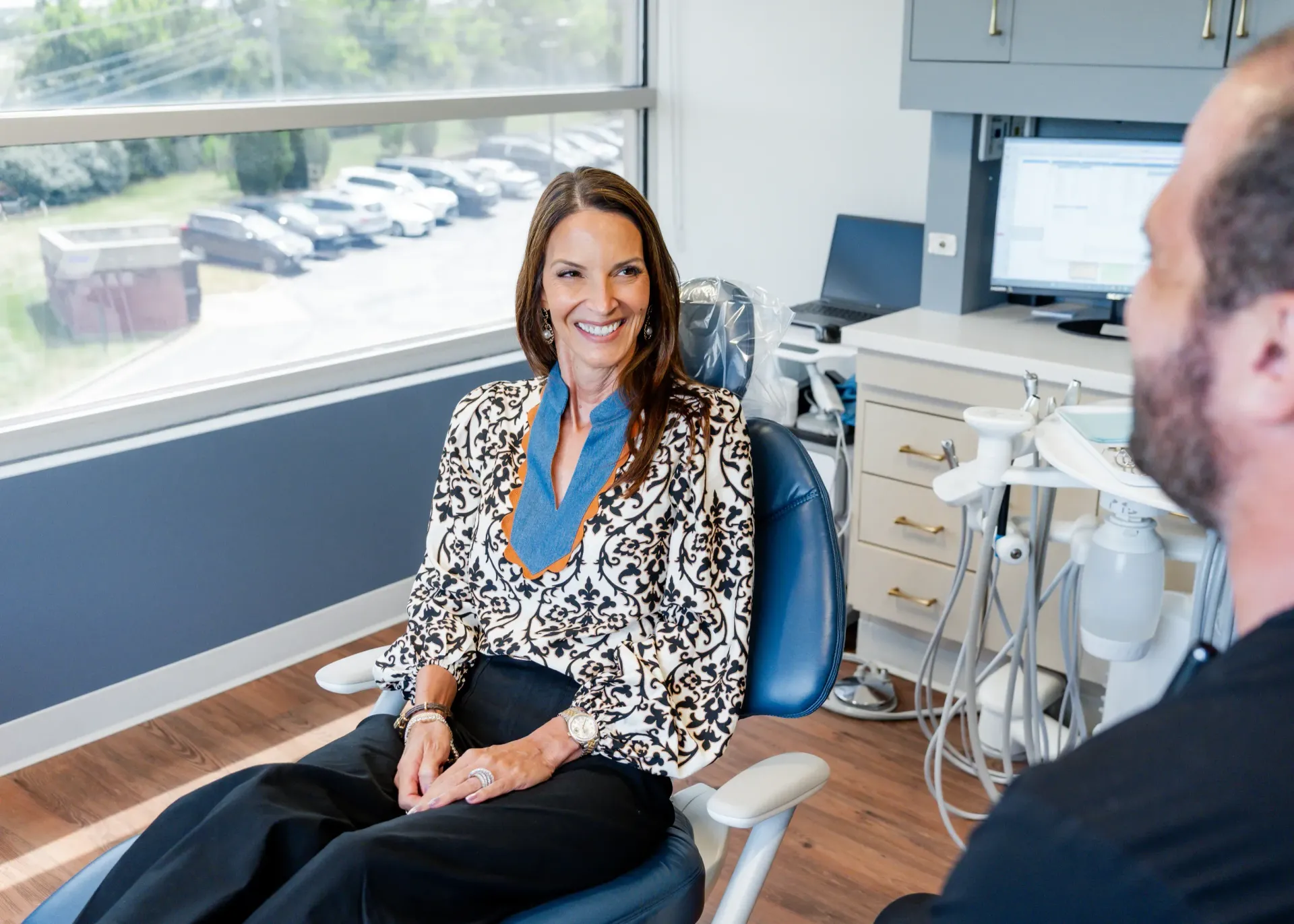 Woman smiles in a dentist chair, facing a dentist in an exam room with a window and equipment.