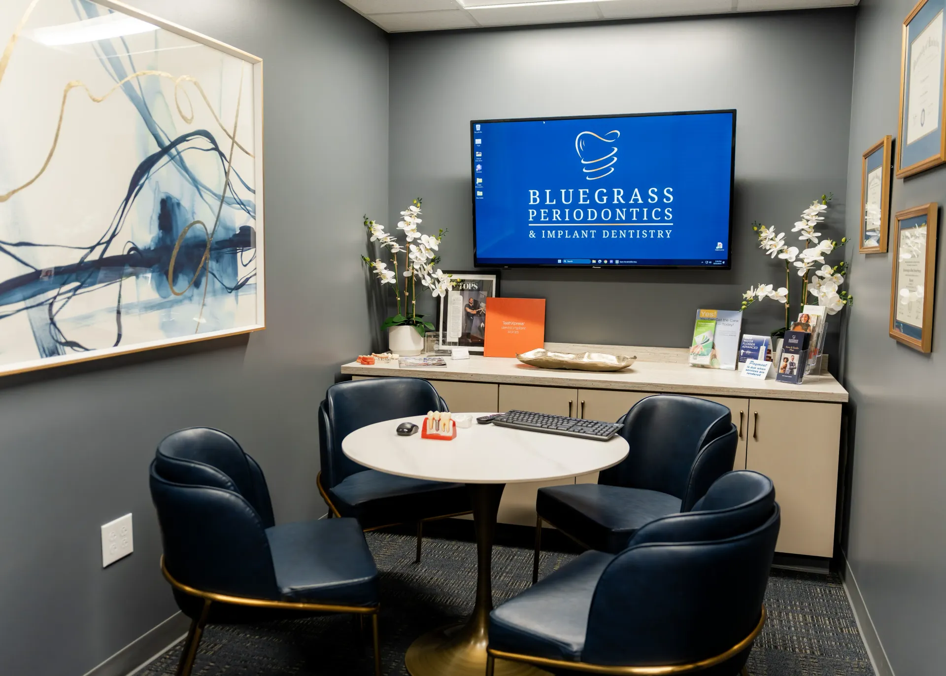 A waiting area with a table, four blue chairs, and a TV displaying a dental office logo.