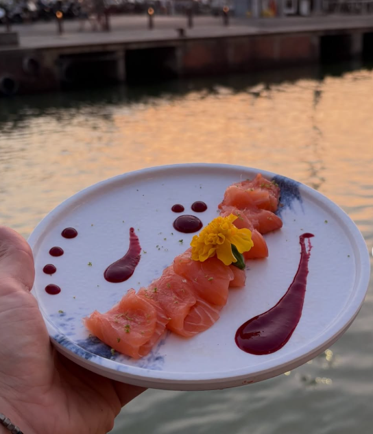 Sashimi di salmone servito con salsa e un fiore giallo, tenuto in mano davanti a un tramonto sul lungomare