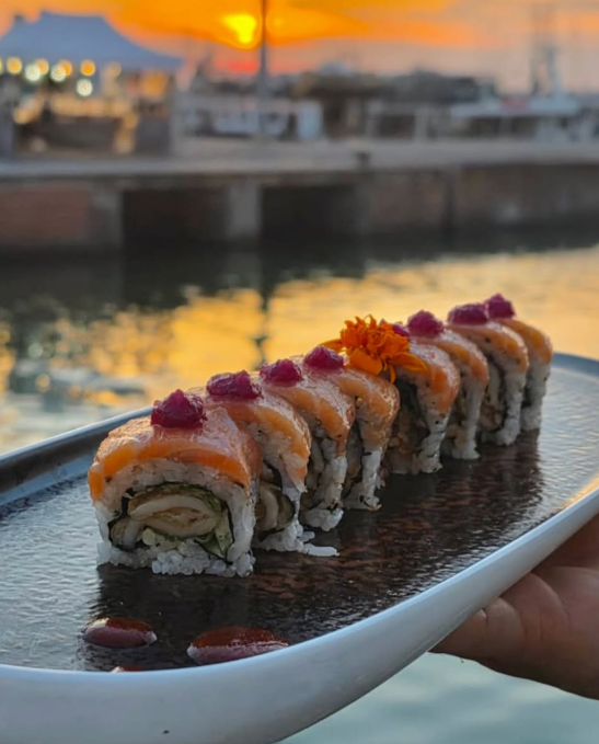 Uramaki conditi con salsa all’arancia e guarnizione viola, serviti su un piatto lungo al tramonto, in riva all’acqua