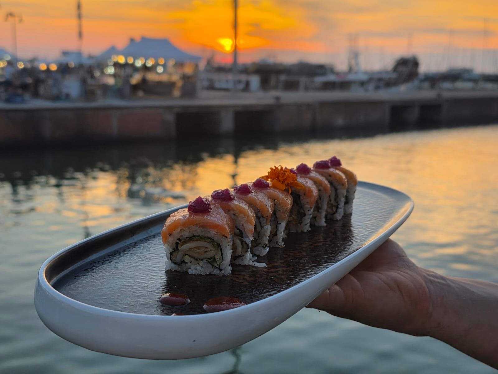 Sushi disposti su un vassoio tenuto sopra l’acqua al tramonto, con un porto sfocato sullo sfondo