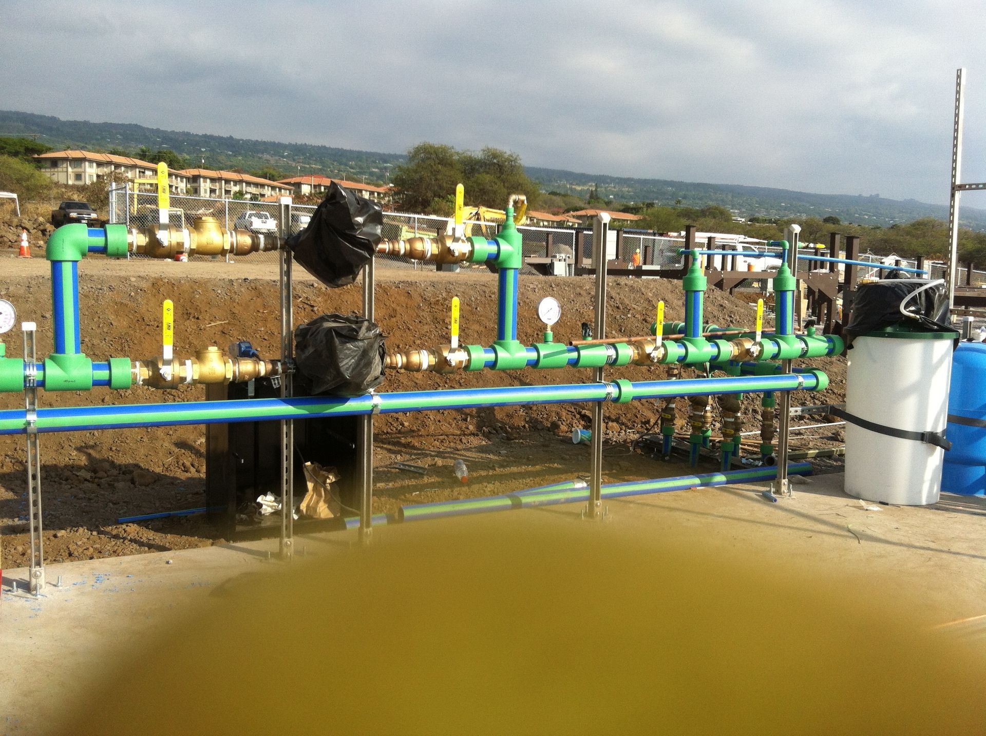 Water treatment pipes and valves over muddy water, with a tank and construction site in the background.