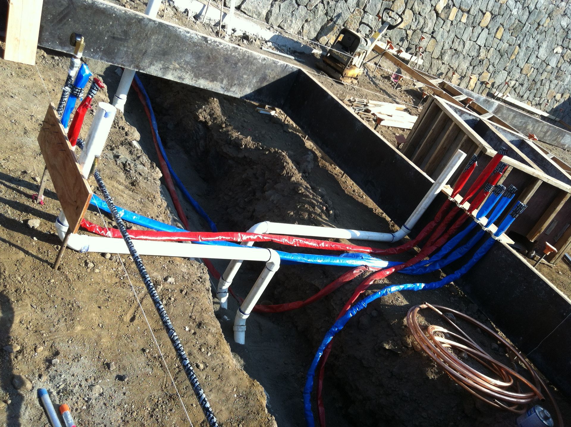 Open trench with red and blue utility pipes and cables beside a stone wall