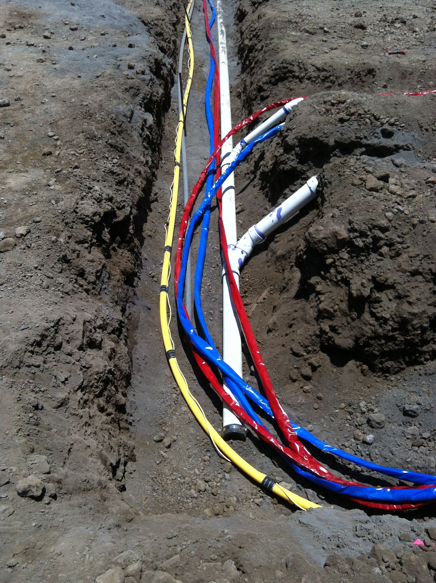 Trench with exposed red, blue, yellow cables and white pipes in the dirt