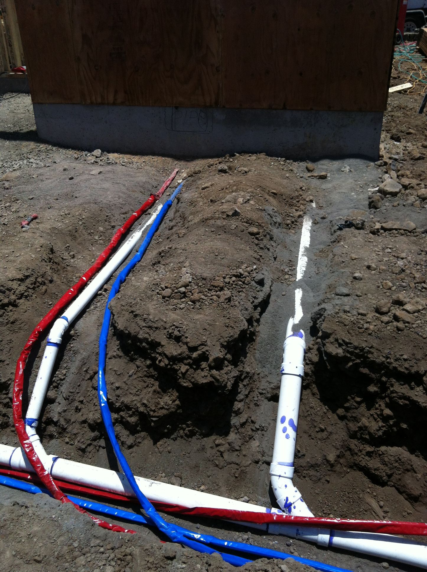 Trenched red, blue, and white pipes running into the ground beside a wooden structure.