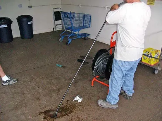 Man holding a long metal tool, possibly unclogging a drain, in a concrete-floored room with shopping carts and a hose reel.