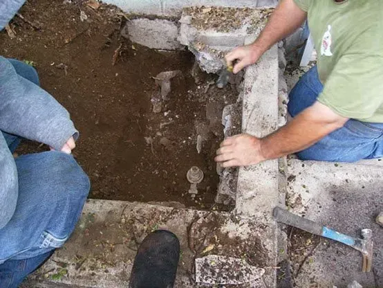 Two people working on the ground near concrete; dirt, tools, and an open area with pipes are visible.