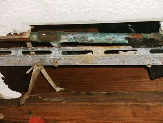 Copper pipes with corrosion, supported by a metal bracket, within a damaged ceiling and wooden framework.