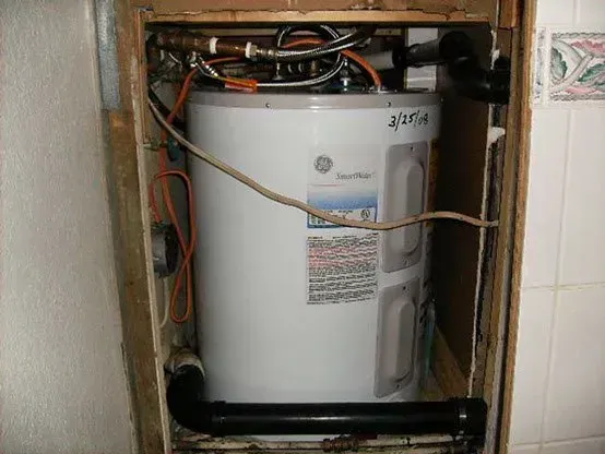 A white GE water heater installed in a wooden framed enclosure, with black plumbing and electrical wiring visible.
