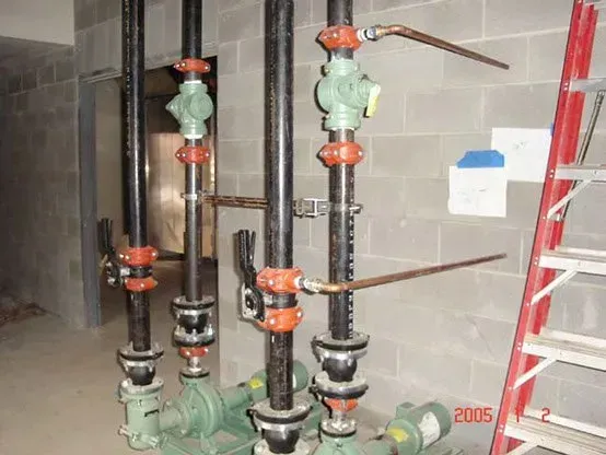 Pipes and valves attached to green pumps against a concrete block wall, next to a red ladder.