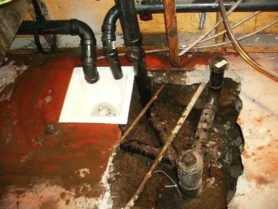 Basement plumbing: Sink and pipes amidst construction debris, red-stained floor, and exposed pipes.