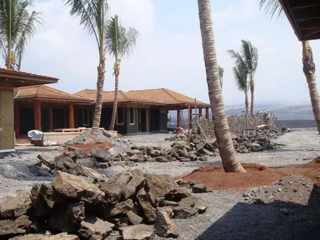 Houses under construction near palm trees and a lava rock fence.