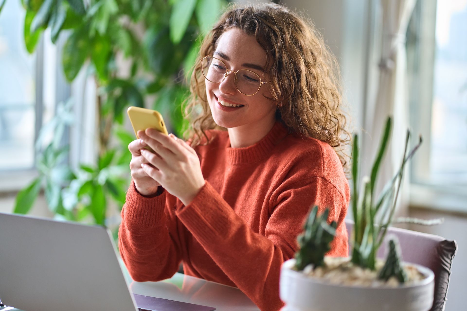 a woman is sitting at a table using a laptop and a cell phone to digitally engage with companies and drive brand affinity