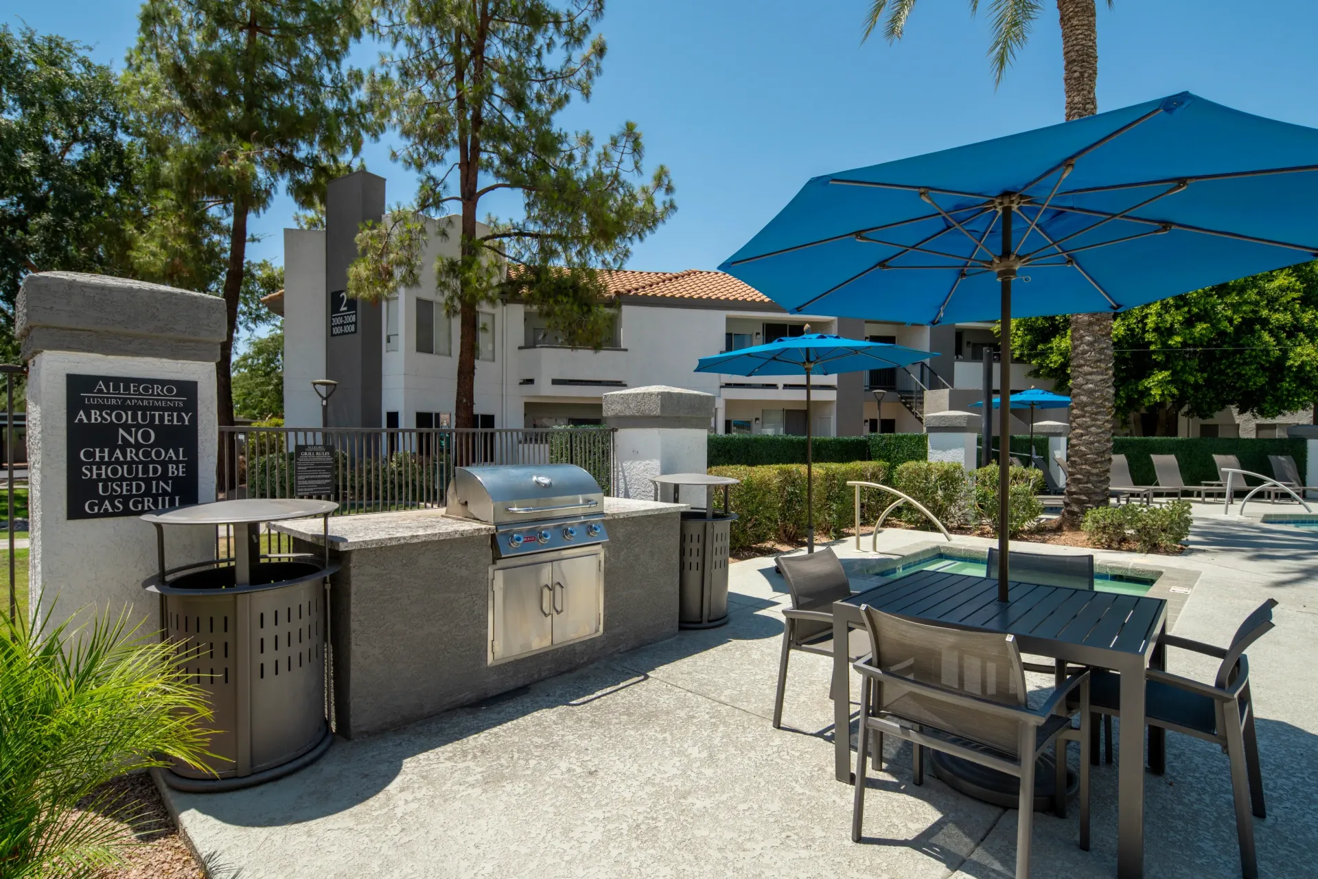 Outdoor grilling station with stainless steel grill, counter space, trash cans, and seating near a swimming pool.
