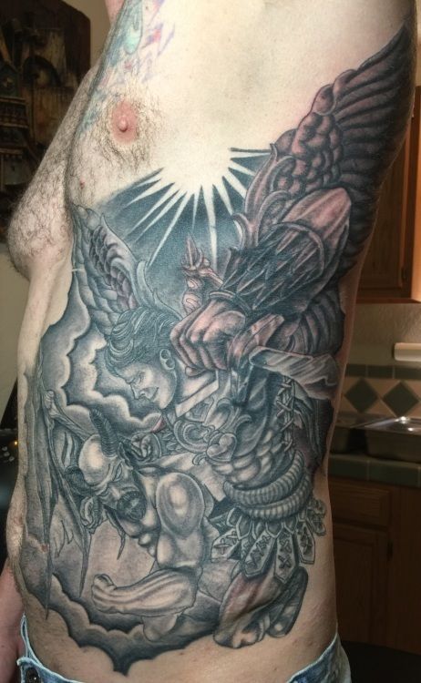 Tattoo on a person's side, depicting a battle scene with an angel, demon, and sunburst in grayscale.